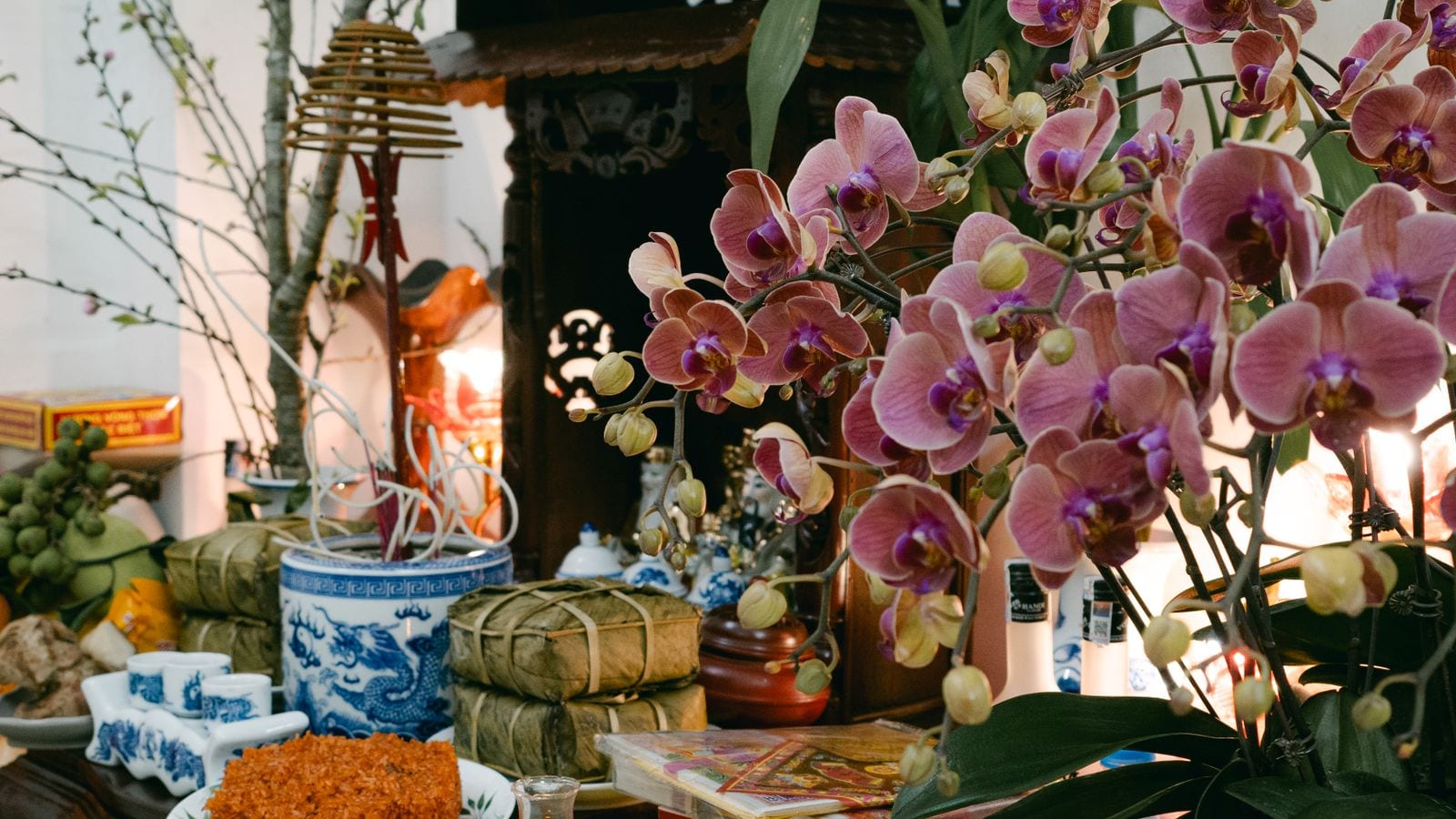 A household altar dressed for Tet: two banh chung tied in leaf, a plate of orange gac sticky rice, blue and white cups, sprays of orchid and peach blossom and a stack of votive papers