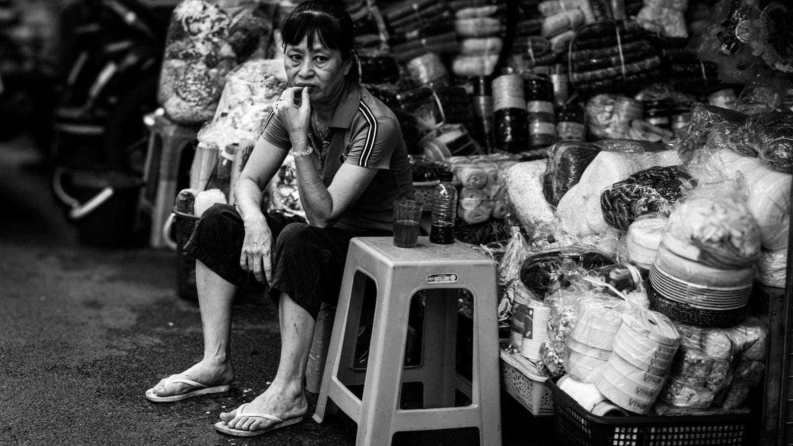 A stallholder sitting on a low plastic stool between stacks of fabric, a glass of tea and the bottle it was poured from on the stool beside her