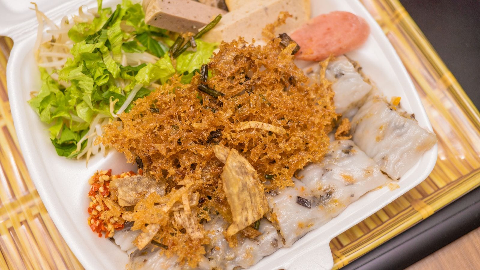 A foam box of banh cuon: rolled rice sheets with minced pork inside, topped with fried shallots and dried pork floss, with sliced gio, salad and chilli beside them