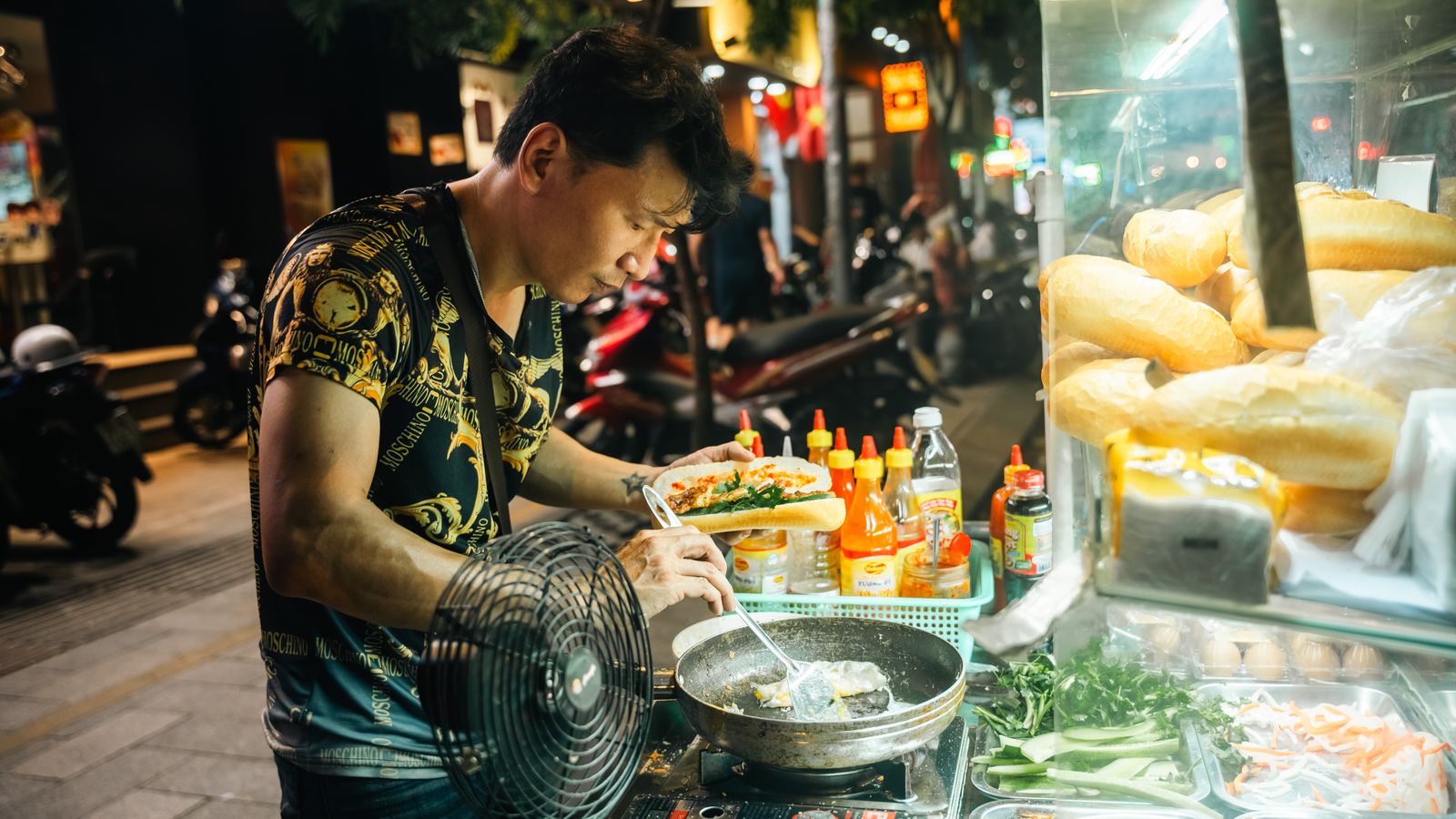 A vendor filling a baguette at a lit banh mi cart at night, sauce bottles and pickled vegetables on the counter