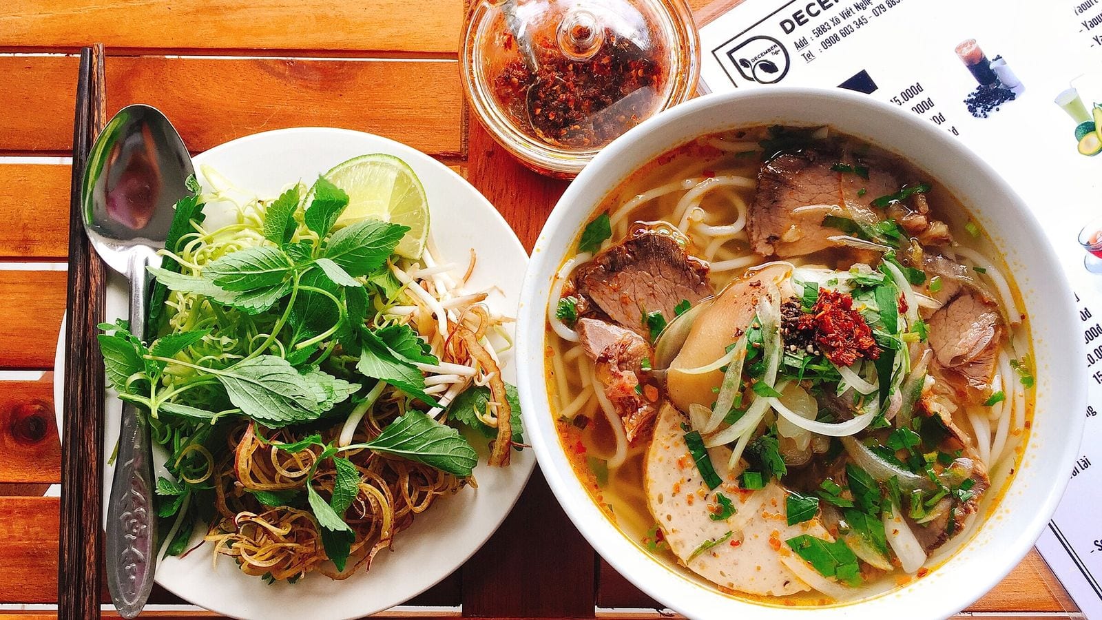 A bowl of bun bo Hue with round noodles, sliced beef, pork loaf and chilli oil, a plate of herbs and banana blossom beside it