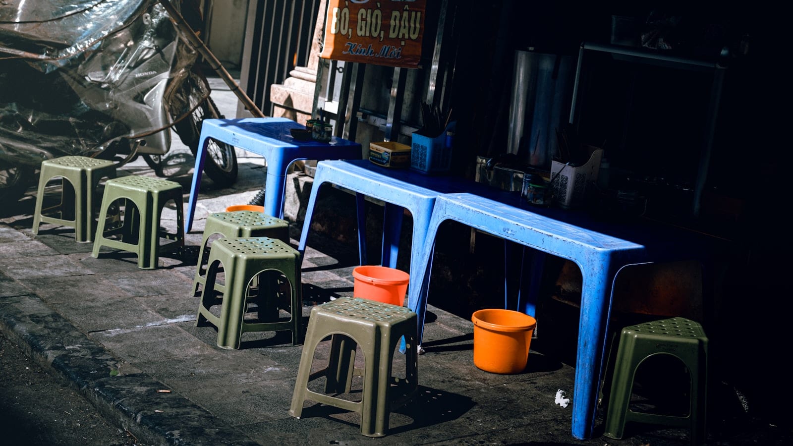 Blue plastic tables and low stools standing empty on a pavement beside a Vietnamese street kitchen
