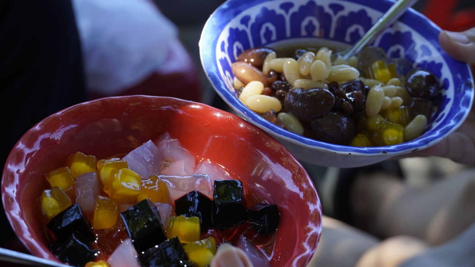 Two bowls of che held up side by side, one with grass jelly and sweetcorn in syrup, the other with white beans and longan