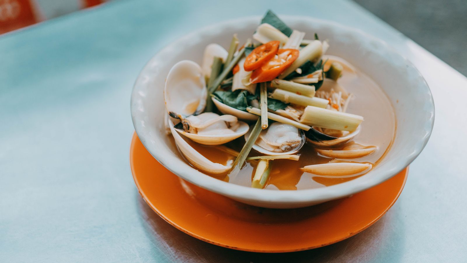 A bowl of clams steamed with lemongrass stalks, chilli and rau ram, sitting on an orange melamine plate on a steel street table