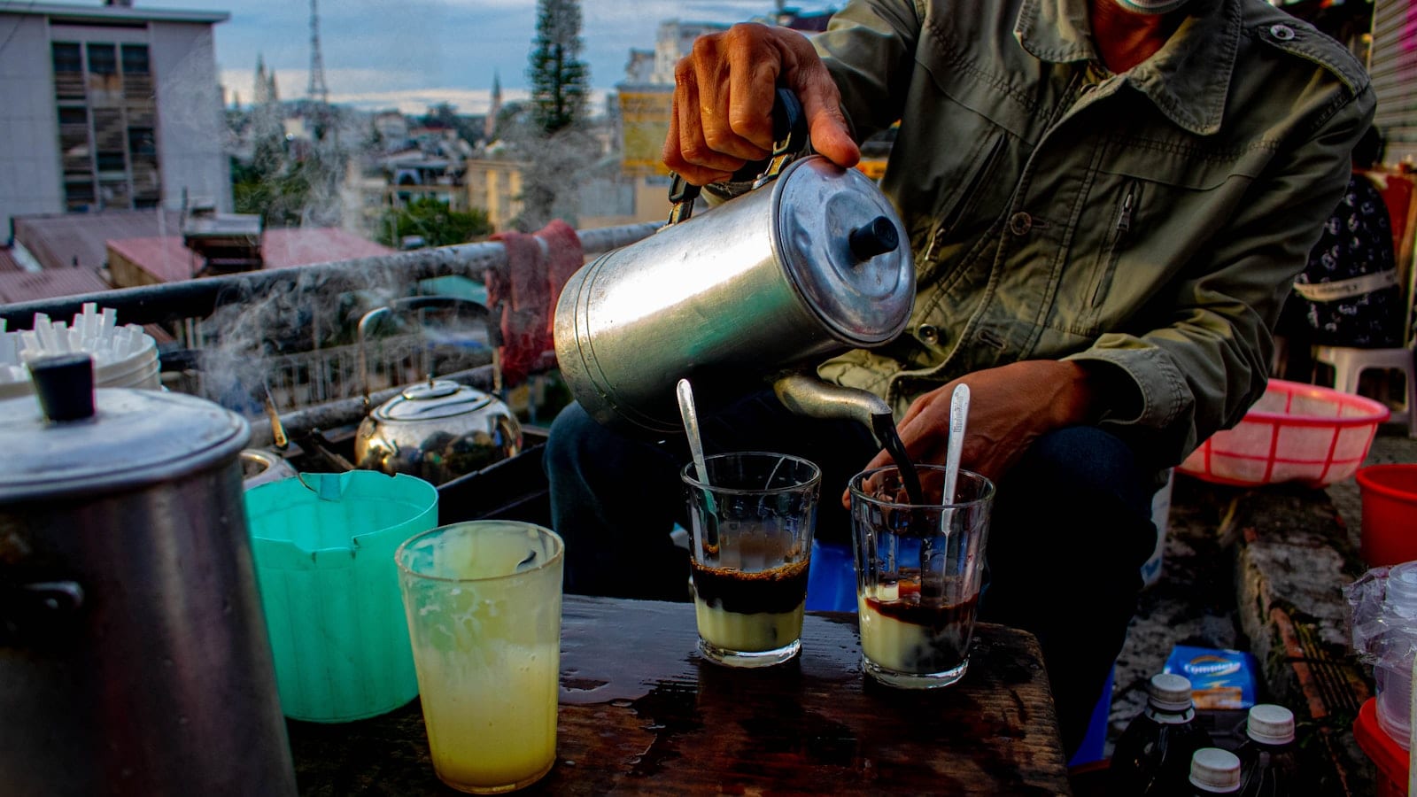 A street vendor pouring from a metal kettle into glasses of coffee, steam rising over rooftops