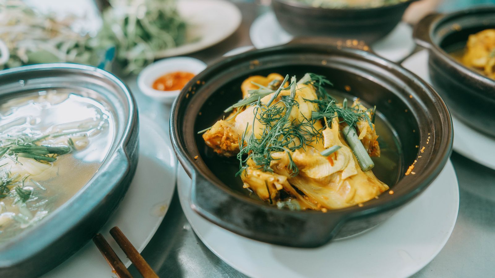 Turmeric-yellow fish under a heap of dill and spring onion in a clay pot, a dish of chilli sauce and herbs beside it