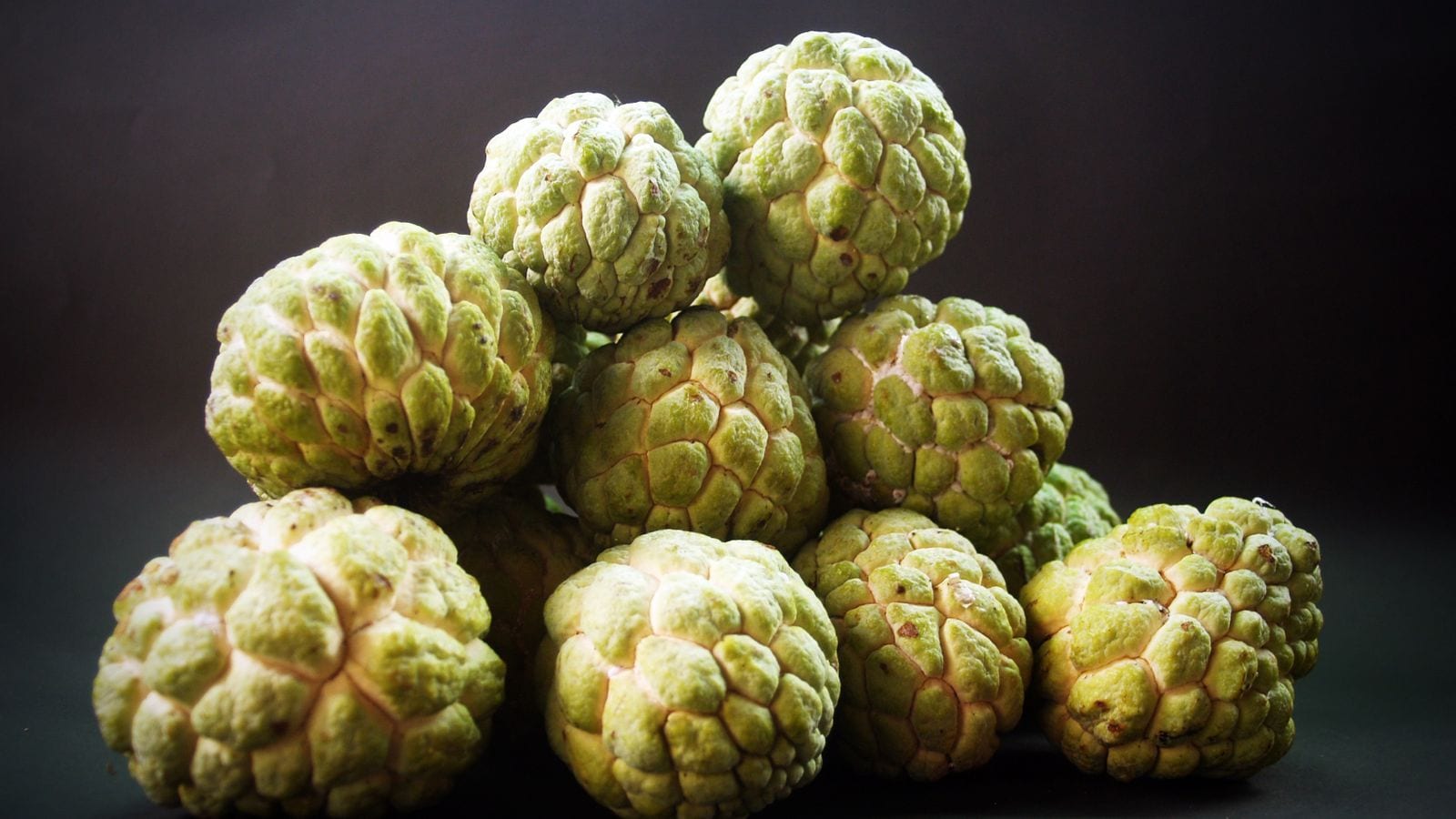 A pile of green custard apples with their scaled skin against a dark background