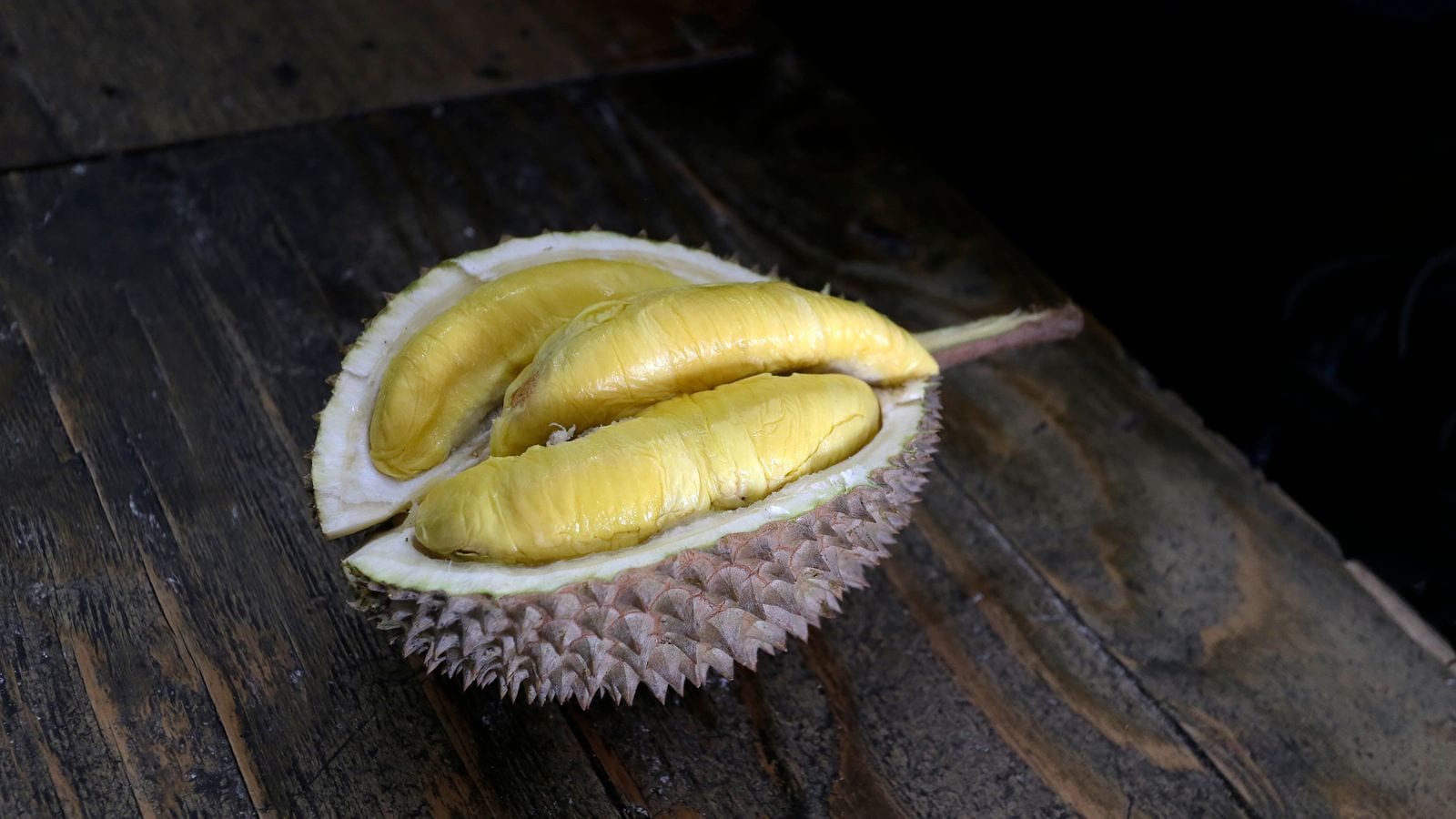 A durian split open on a wooden table, four pale yellow lobes of flesh inside the spiked shell
