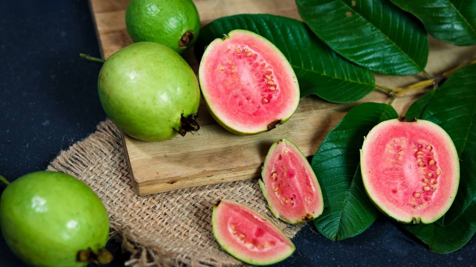 Whole green guavas beside halves showing pink flesh and seeds, guava leaves around them