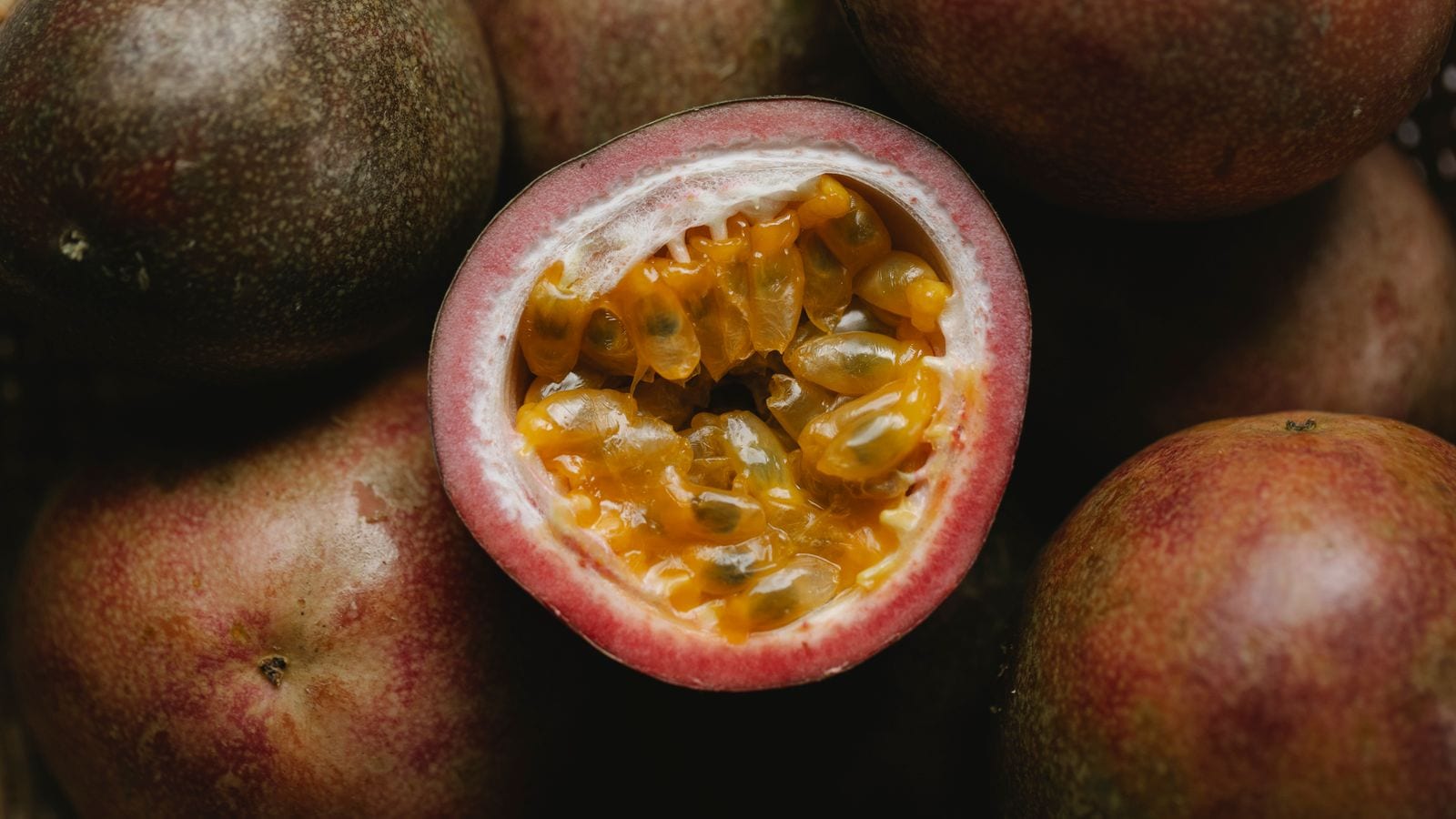 A passion fruit cut in half showing orange pulp and seeds, whole fruits around it
