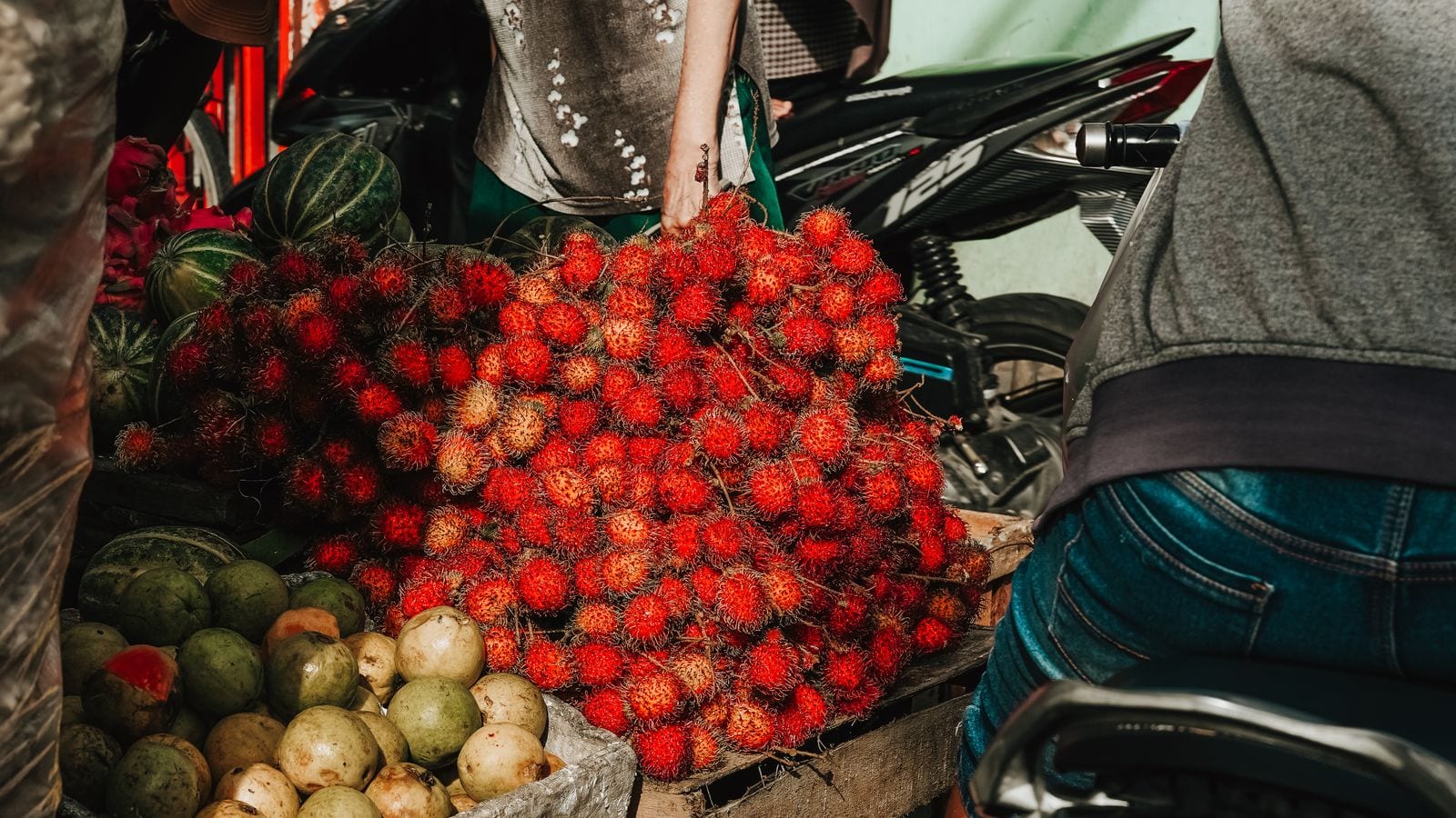 A heap of red rambutan on a market stall beside guavas and watermelons, a scooter parked behind