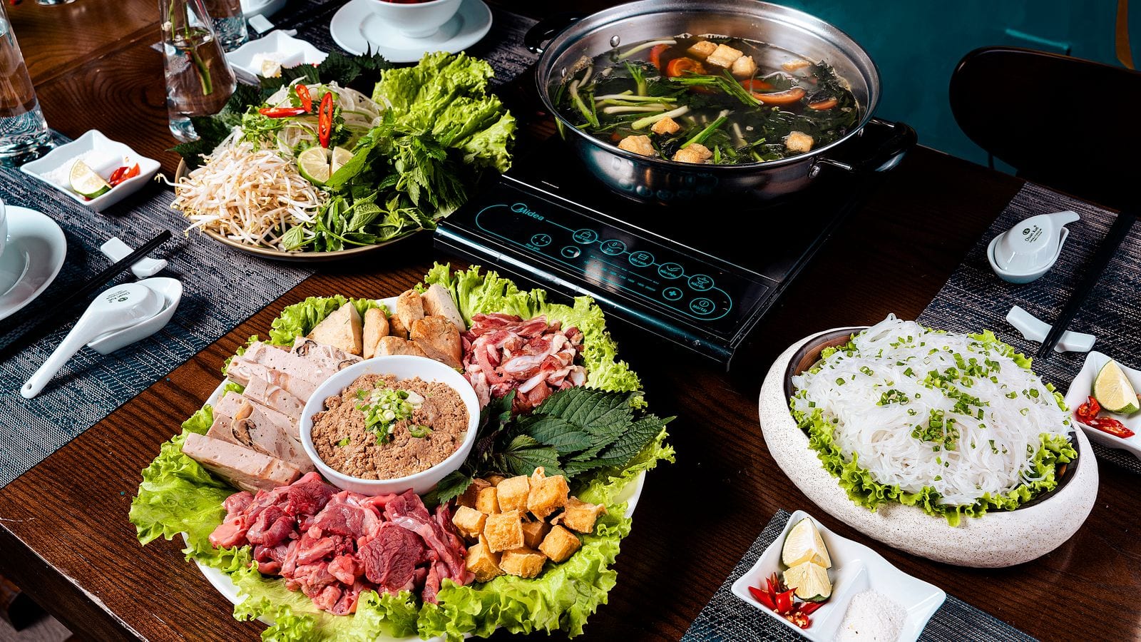 A lau table: a steel pot simmering on an induction plate, with raw beef, pork sausage, fried tofu, crab paste, perilla leaves and a bowl of bun laid out around it