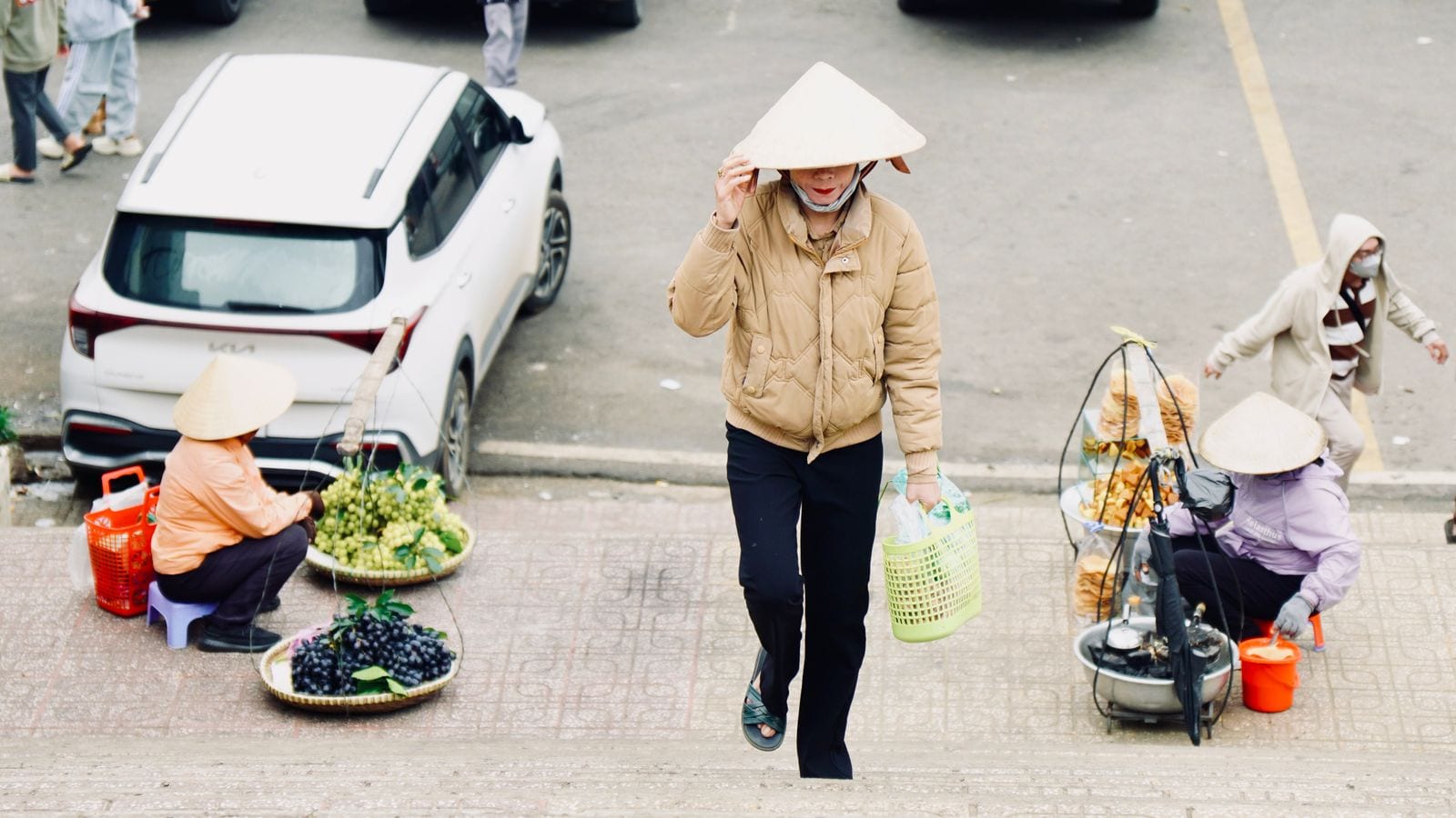 Two vendors in conical hats squatting on a pavement beside flat bamboo trays of grapes and longan, shoulder poles laid on the kerb behind them