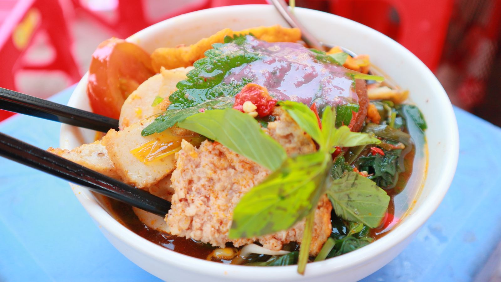 A bowl of bun rieu on a blue plastic table: crab patty, tomato, tofu puffs and herbs in a red-orange broth