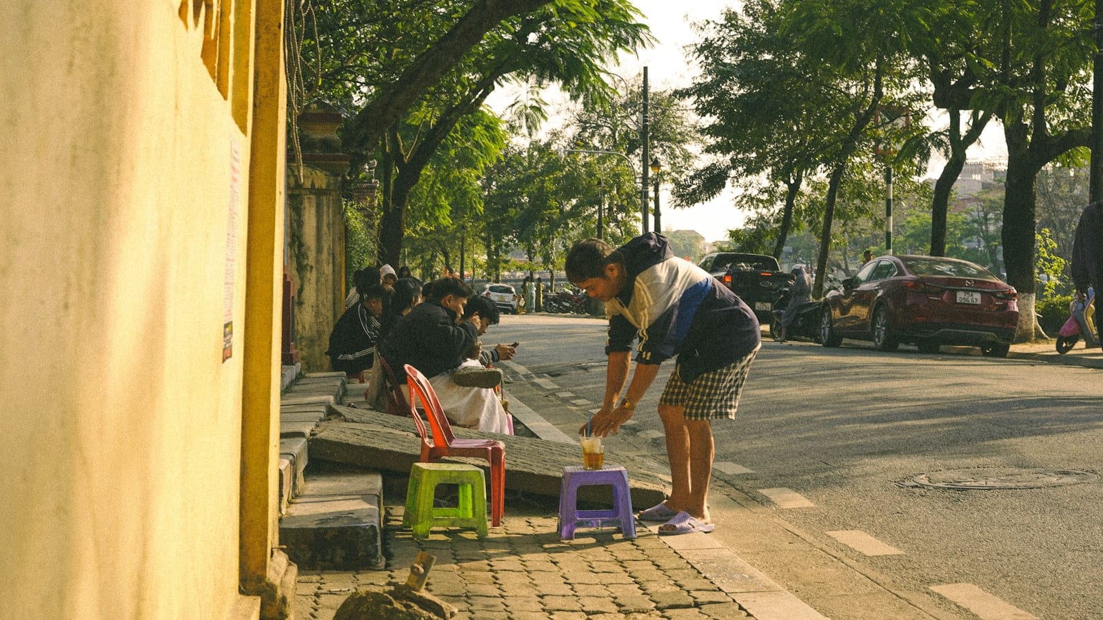 Low plastic stools set out on a Vietnamese pavement in morning light, a glass of iced tea being placed on one and customers sitting along the kerb