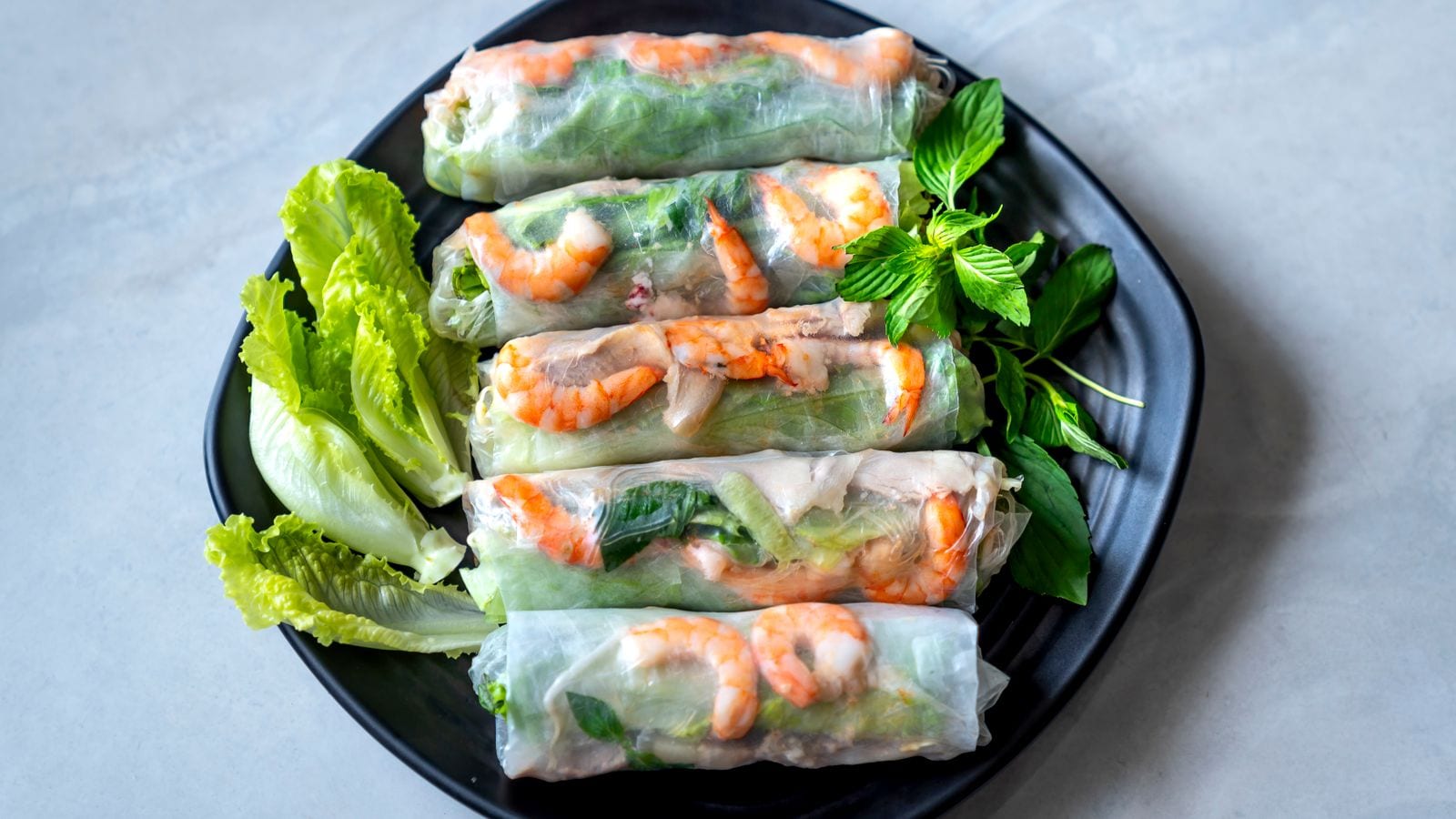 Five goi cuon on a dark plate, prawn and pork visible through the rice paper, with lettuce and Thai basil beside them