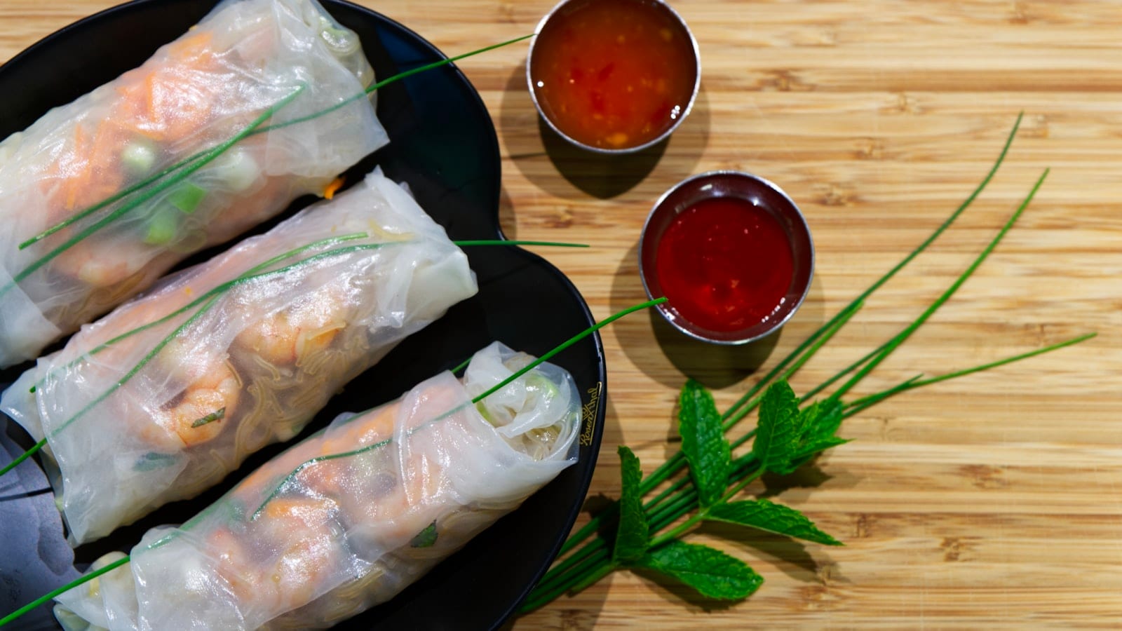Fresh rice paper rolls on a black plate with two dipping sauces and mint