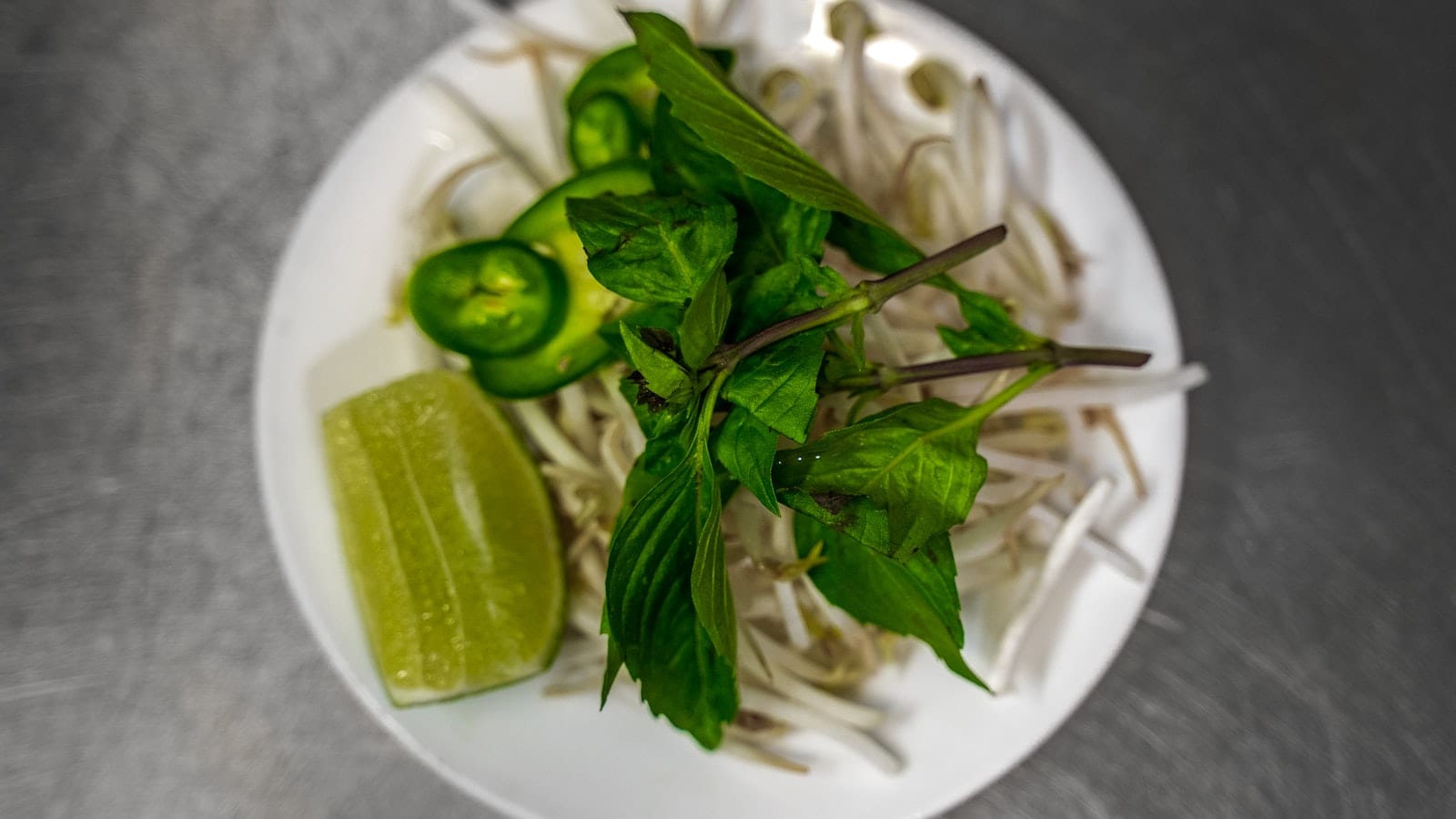 Plate of raw bean sprouts, Thai basil sprigs, sliced green chilli and a lime wedge, seen from above