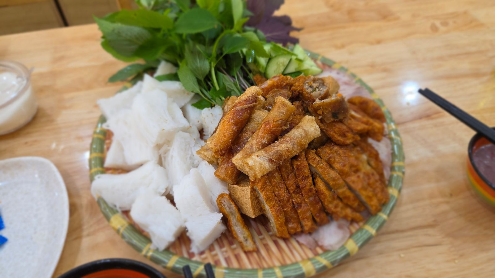A bamboo tray of sliced fried pork and rice sheets with a pile of raw herbs beside it — perilla, Vietnamese balm and coriander, stems and all