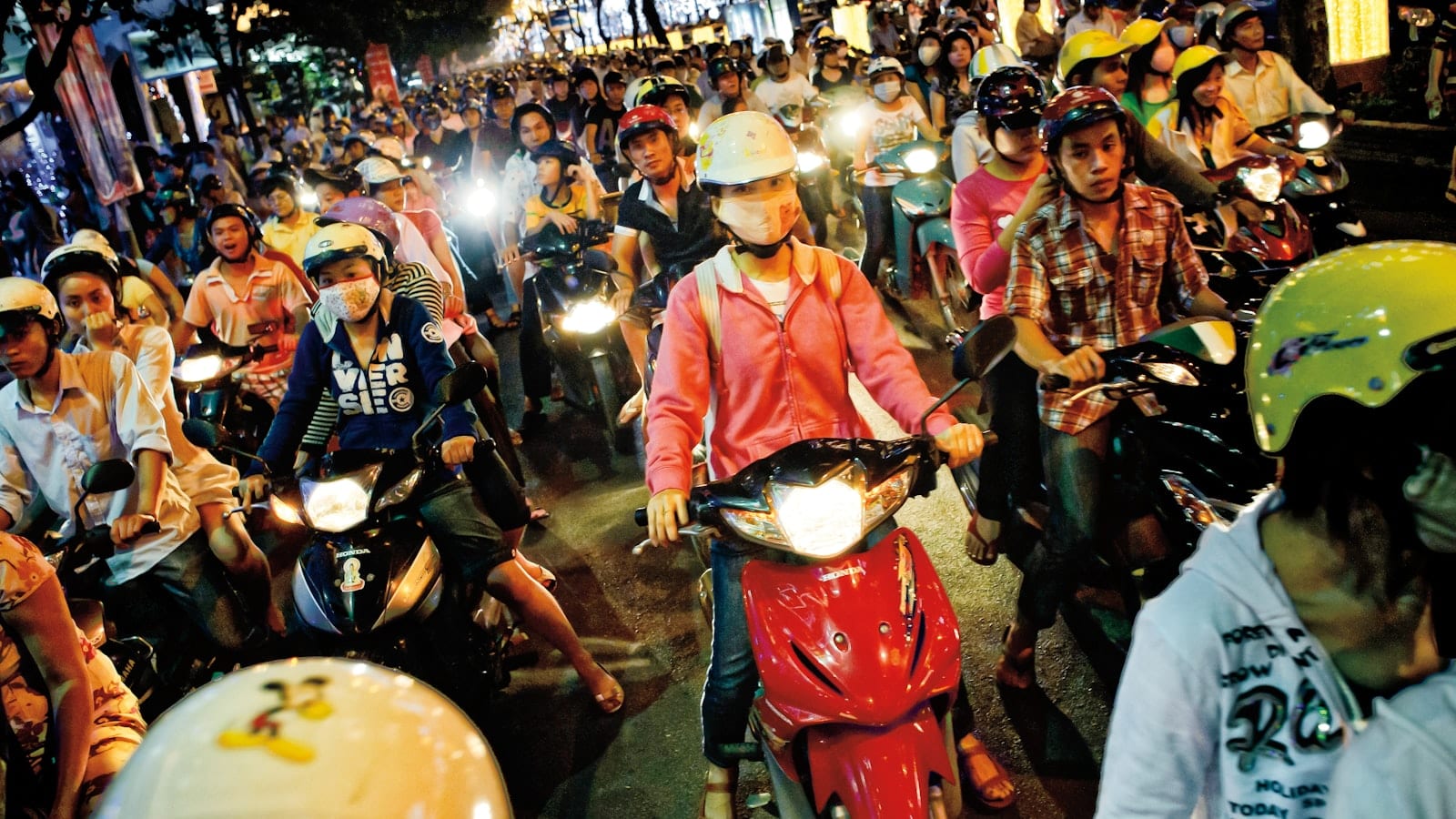A dense crowd of scooter riders in helmets stopped at a junction at night, headlights on