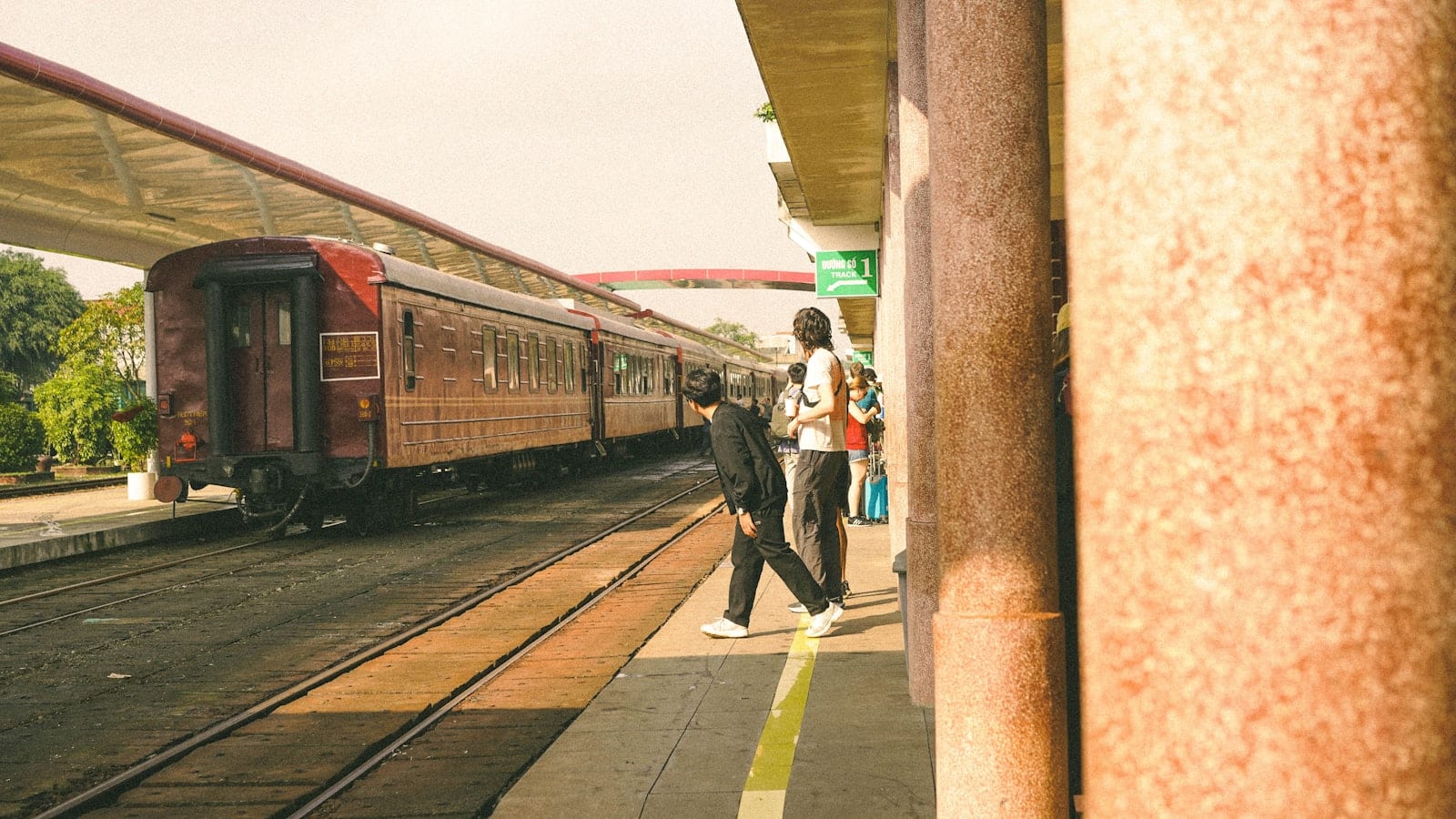 Maroon carriages at a Vietnamese station platform, passengers waiting under a Track 1 sign