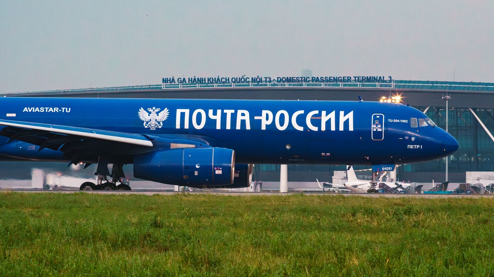 A cargo aircraft taxiing past a terminal lettered NHA GA HANH KHACH QUOC NOI T3 - DOMESTIC PASSENGER TERMINAL 3, with airliners parked at the gates behind it