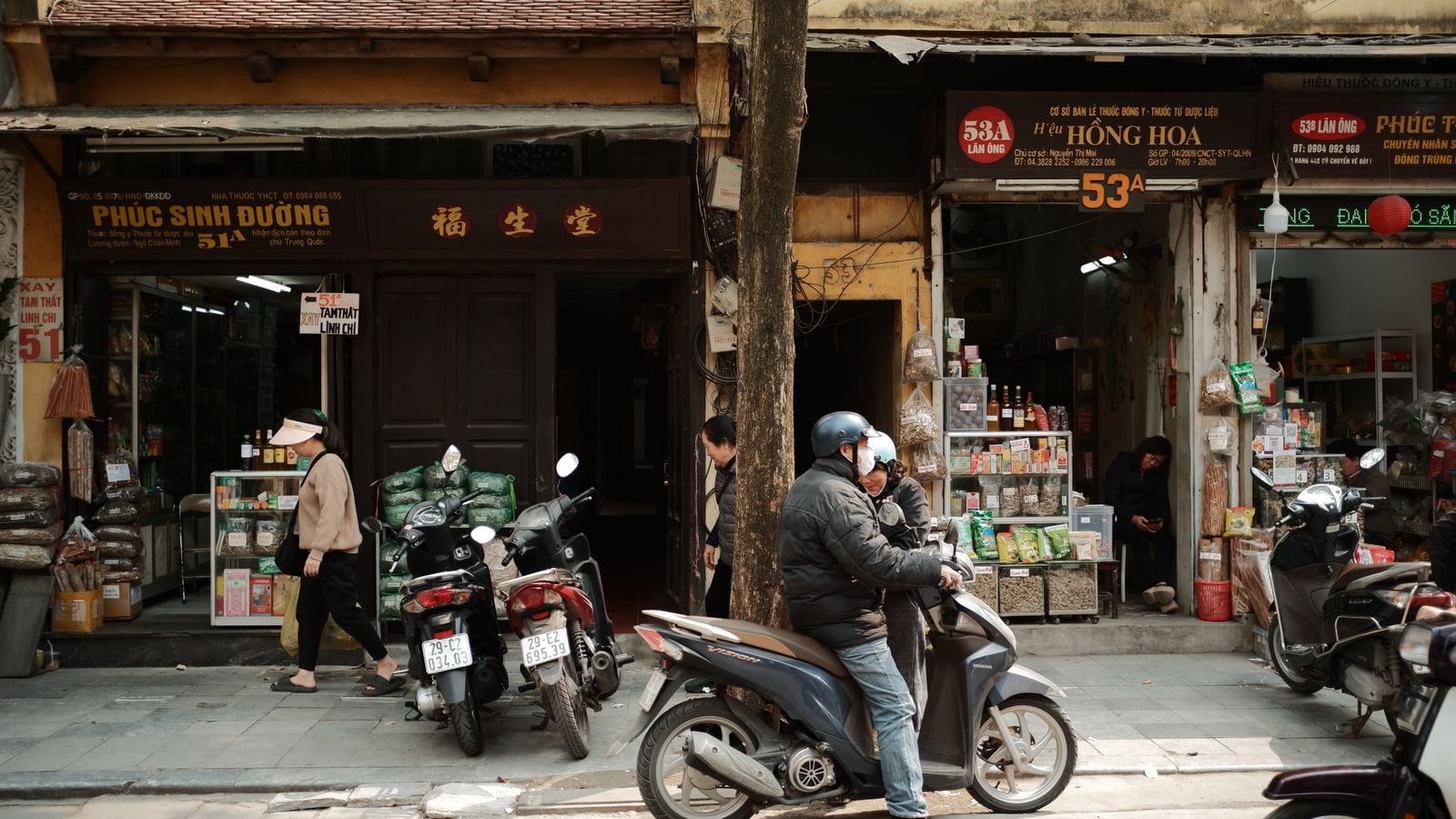Two herbal medicine shopfronts side by side on Lan Ong street, sacks and jars of dried ingredients out on the pavement and a rider stopped between them