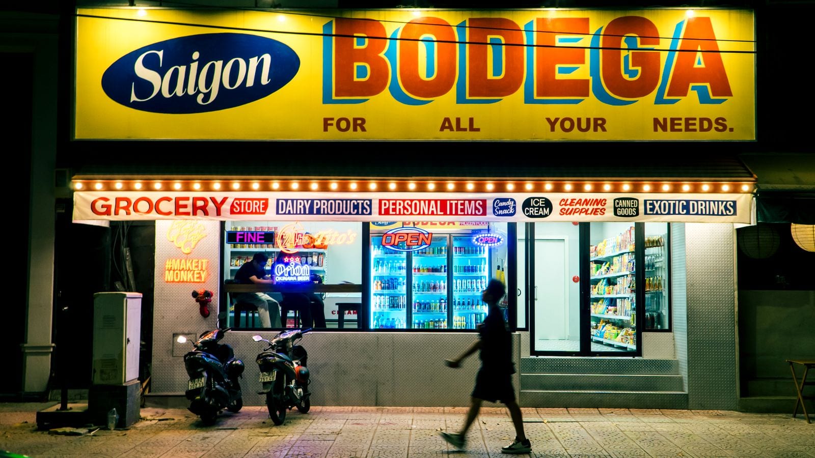 An independent grocery lit up at night under a hand-painted Saigon Bodega sign, chilled drinks cabinets facing the street and two scooters parked at the kerb