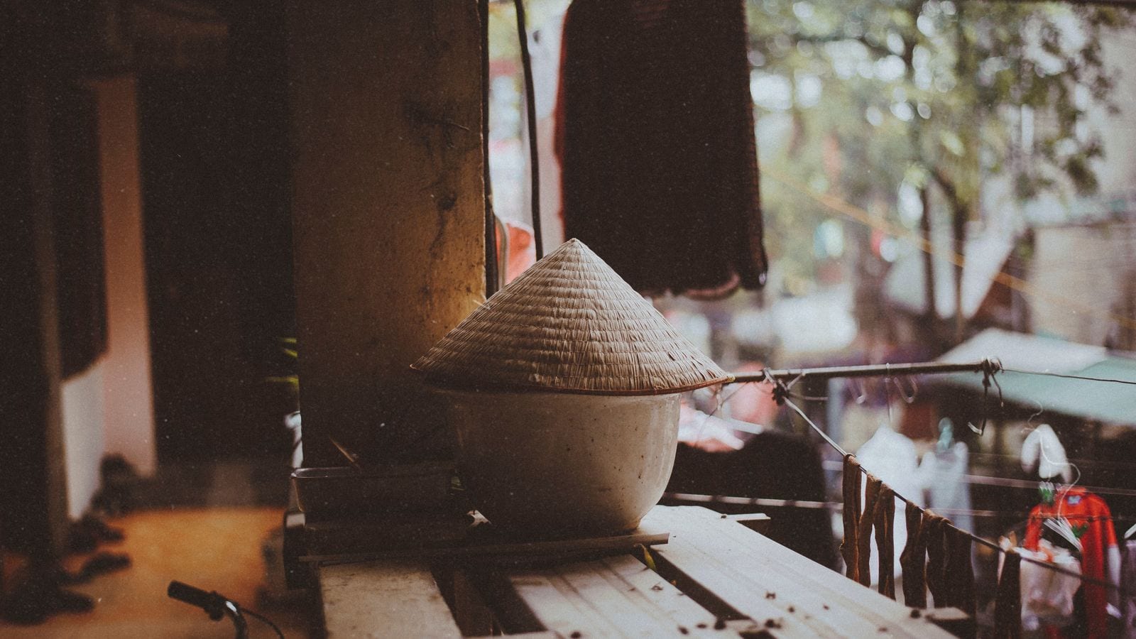 A conical hat resting upside down on a bowl by an open shopfront, with washing pegged along a line strung across the lane outside