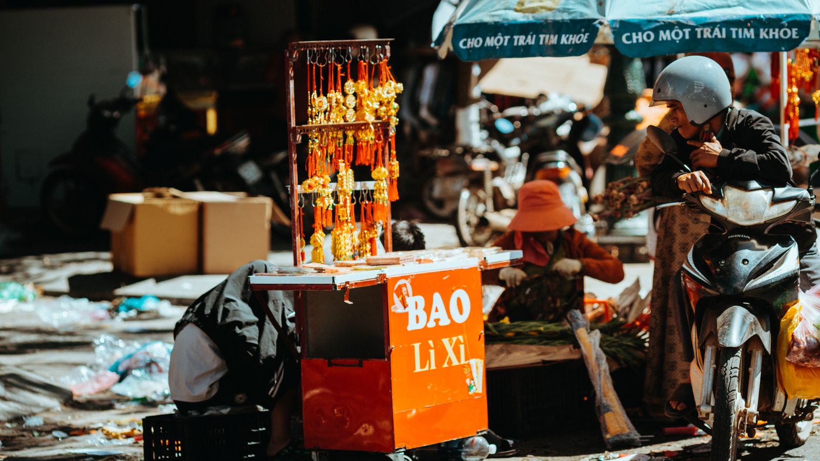 A street cart selling red envelopes marked BAO LI XI, gold charms hung on a rack above it, a rider pulled up alongside and a market umbrella behind
