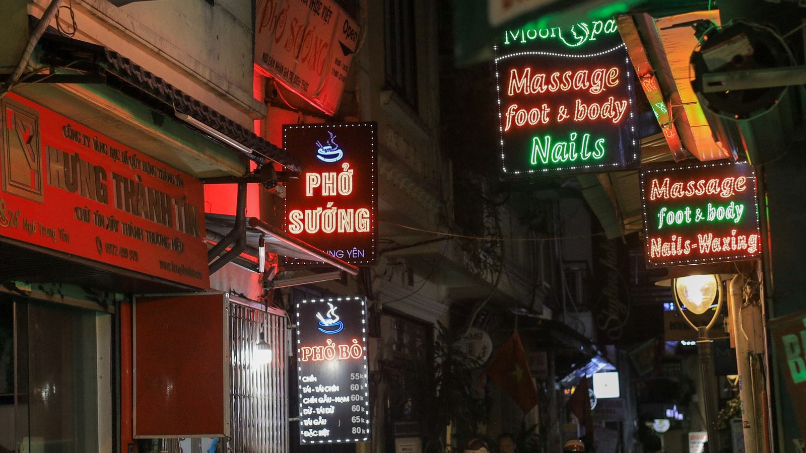 Lit signs stacked over a narrow lane at night: two reading Massage foot and body, Nails and Waxing, a pho shop sign beside them and a price list in thousands below