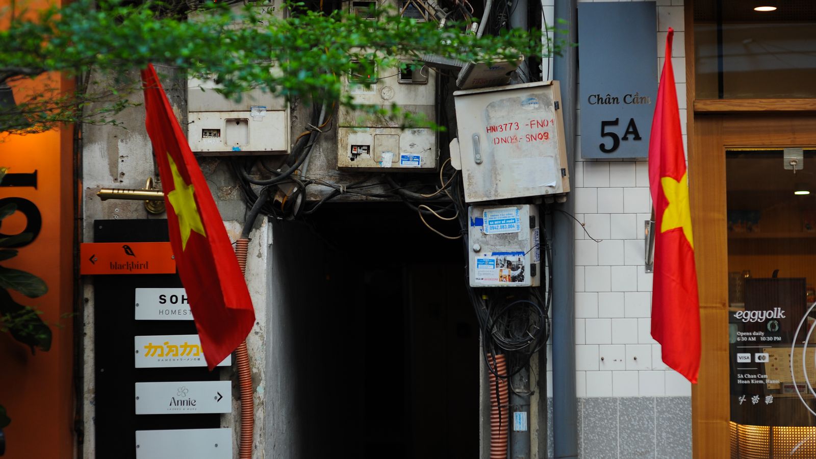 The mouth of an alley at 5A Chan Cam: the number plate on the tiled wall, a stack of business signs for a homestay, a ramen bar and a nail salon beside the dark entrance, and two national flags either side