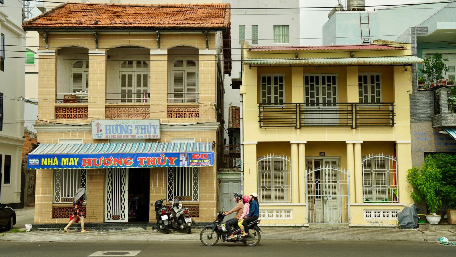 A tailor shop under a yellow and blue awning reading NHA MAY HUONG THUY, in the ground floor of an ochre colonial villa with two scooters parked outside