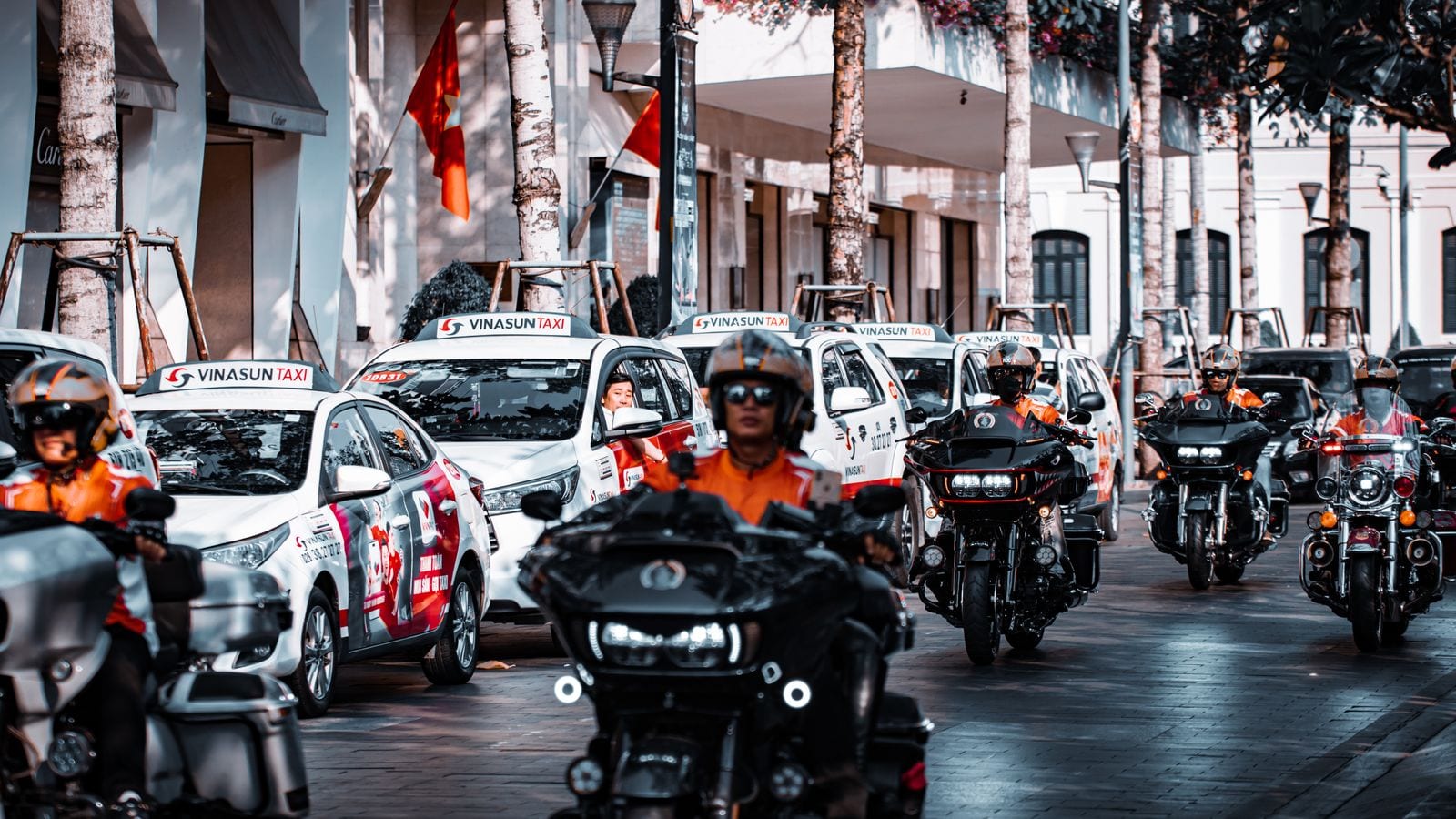 A rank of white Vinasun taxis waiting nose to tail along a city street, motorbike outriders passing in front of them and national flags hanging from the buildings
