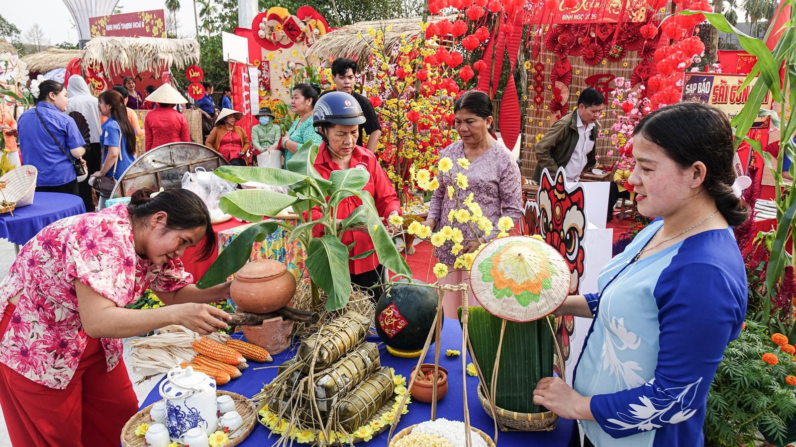 A Tet market stall: bundles of banh tet tied with straw, yellow apricot blossom, red lanterns and a paper lion head behind the table