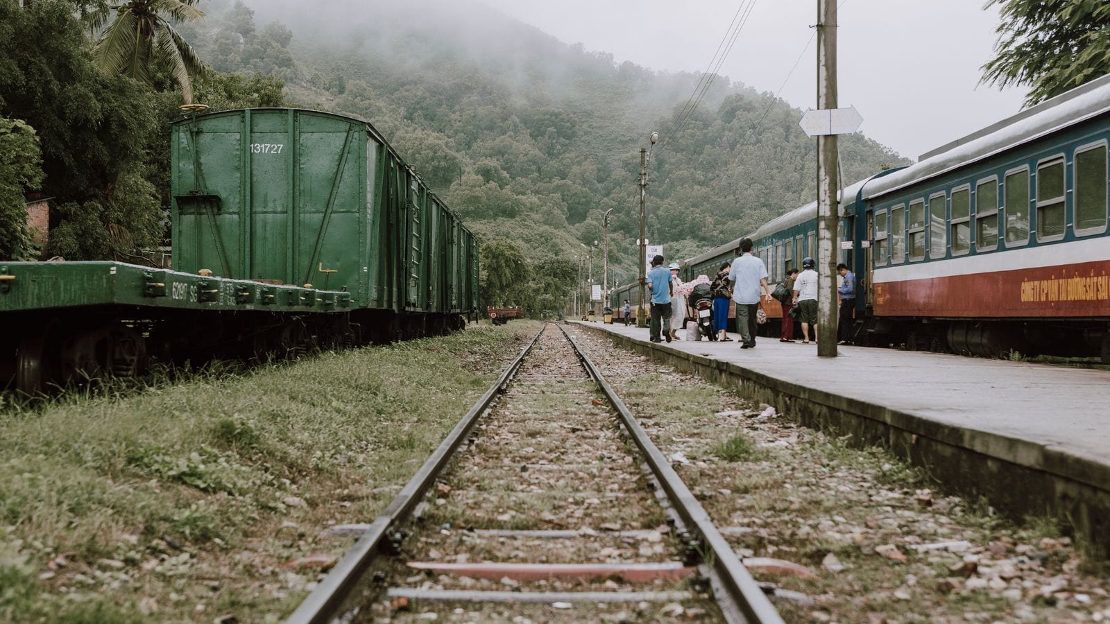 Passengers walking along the platform of a small railway halt under forested hills, a green goods wagon standing on the far track and the carriage lettered for the Saigon railway company
