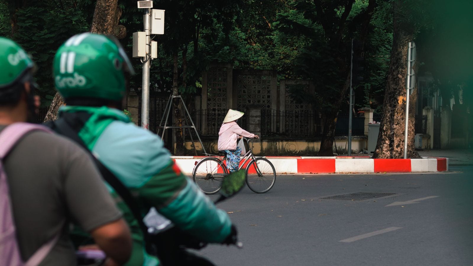 Two riders in green Grab helmets and jackets waiting at a junction, a woman in a conical hat pushing a bicycle across the road in front of them