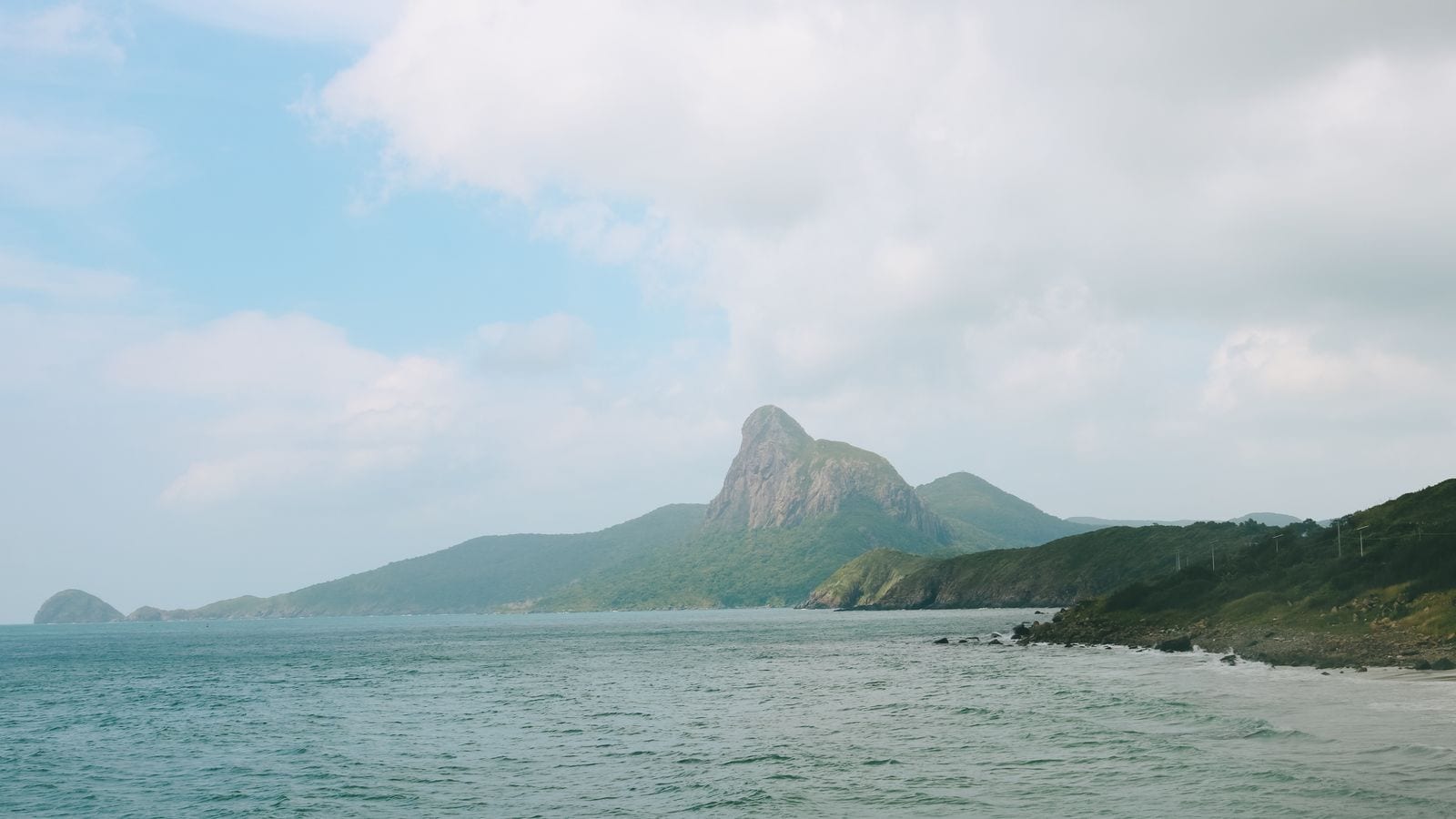 An empty island coast under high cloud, a bare rock peak standing above the forested headland and not a building in sight along the shore