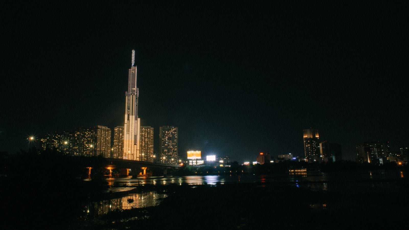 A single lit tower standing far above everything around it on a dark river bank, the rest of the city low and scattered along the water