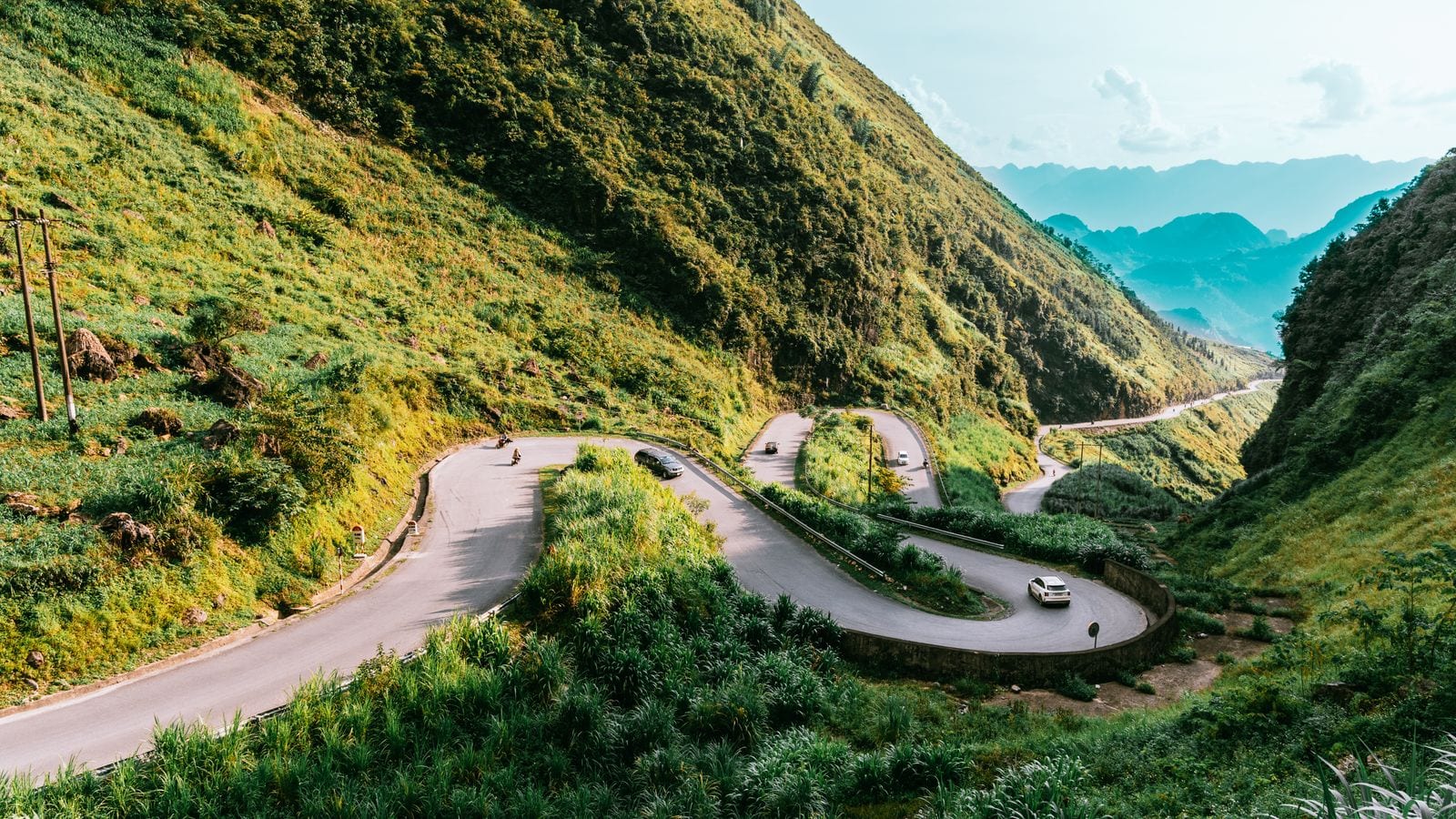 A mountain road folding back on itself three times down a green slope, two cars and a motorbike strung out along the bends and blue ridges behind