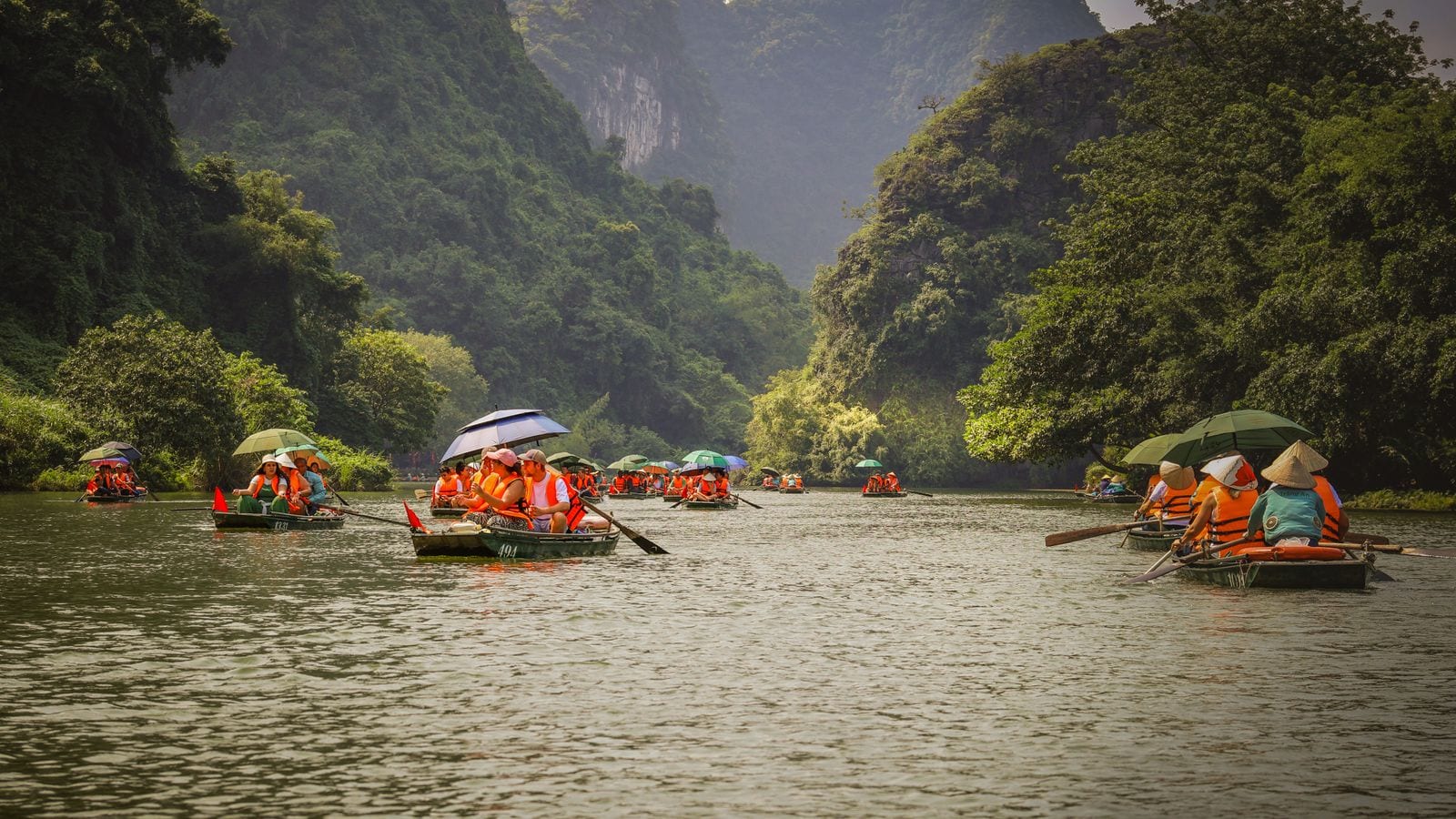 Numbered rowing boats under bright umbrellas on a green river between wooded limestone hills, rowers in conical hats working the oars with their feet