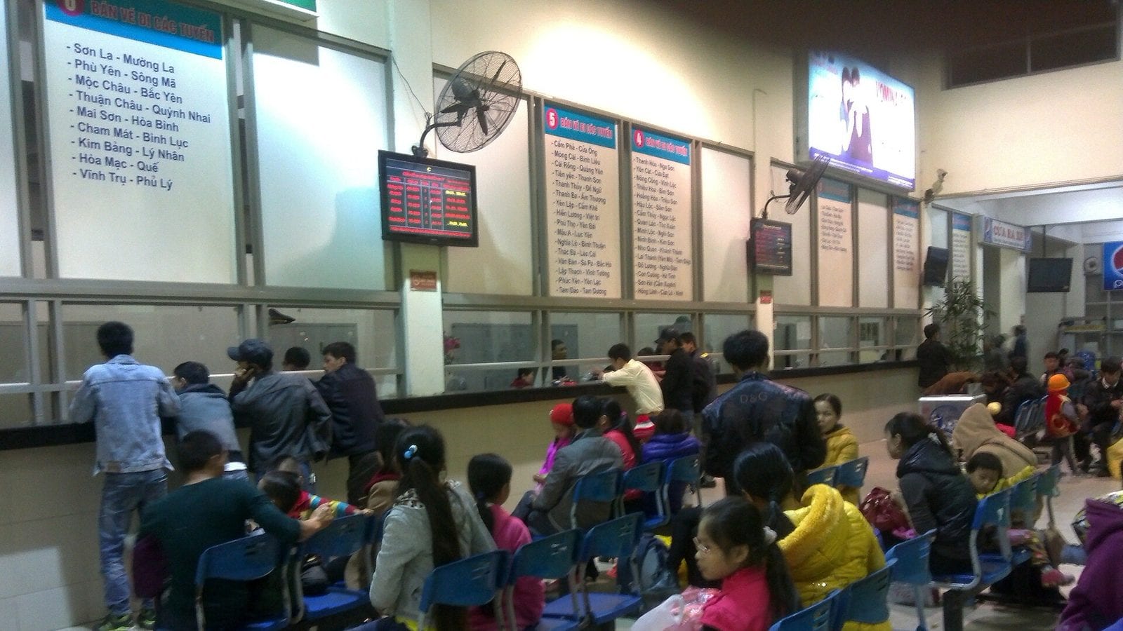 The ticket hall of a bus terminal, route lists painted above every window — Son La, Moc Chau, Mai Son, Phu Ly — a red LED departure board between them and rows of blue seats filled with waiting passengers