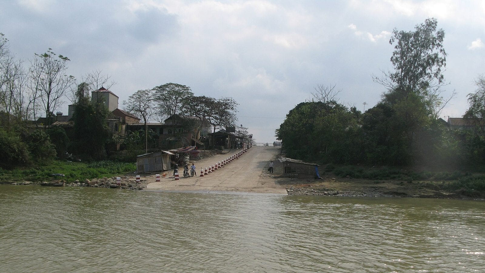 A concrete slipway running straight down into a brown river from a village street, roadside stalls either side of it and a man walking down to the water