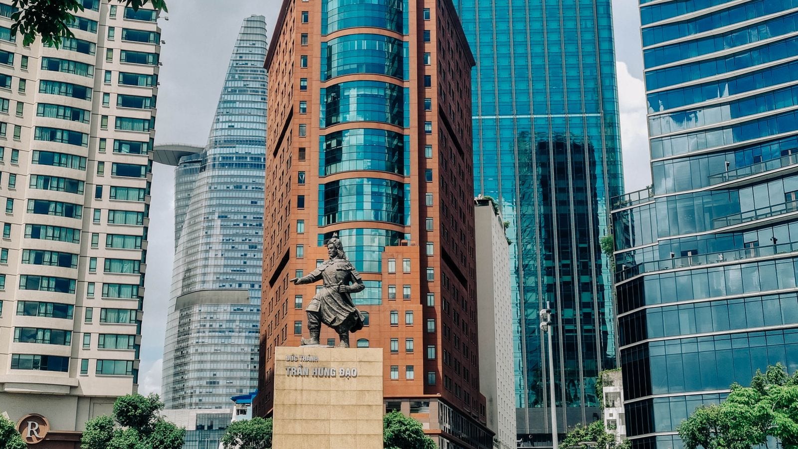 A bronze general on a stone plinth in the middle of a city square, glass and brick towers rising close behind him on three sides