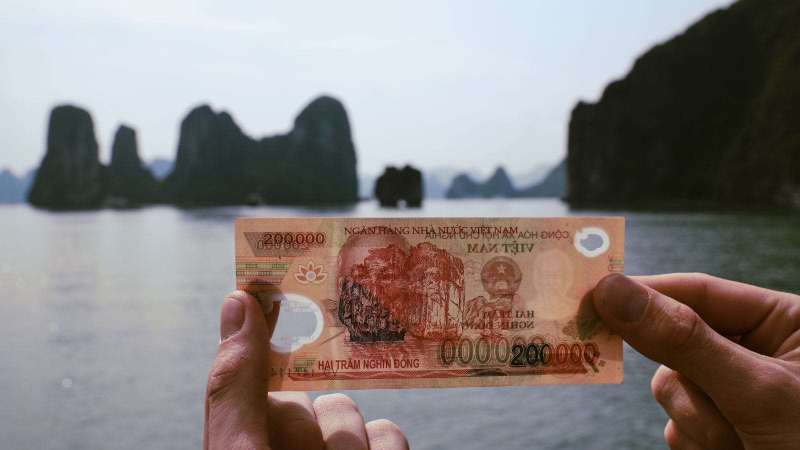 A 200,000 dong note held up in two hands against the bay it is engraved with, the same limestone stacks behind the paper and on it