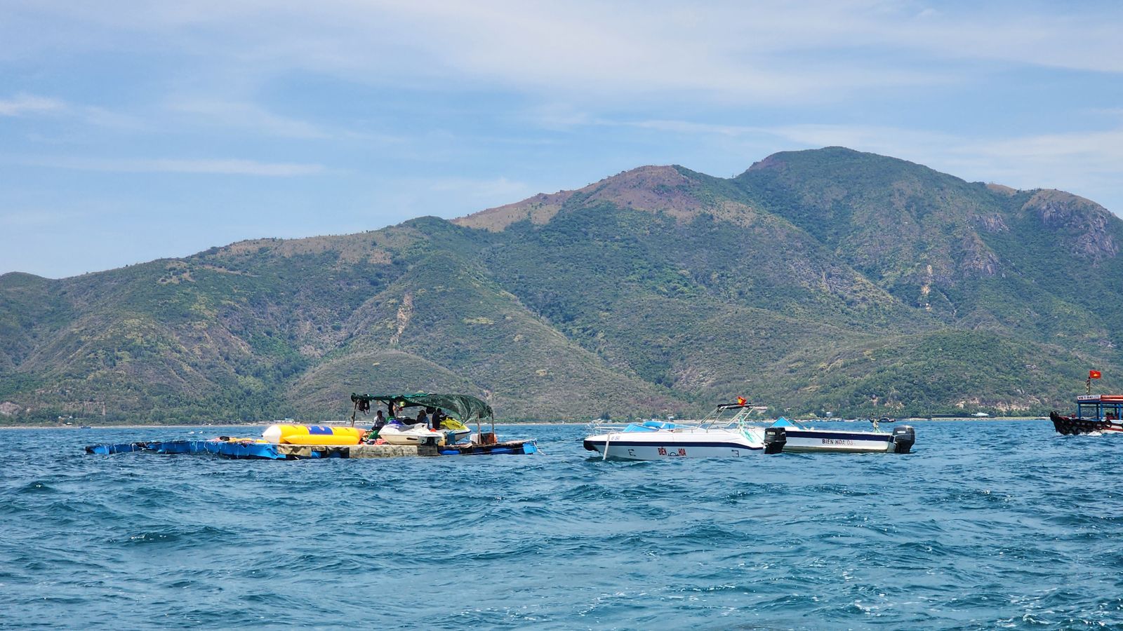 Two speedboats and a floating platform of banana boats and jet skis moored off a scrubby island, a national flag on a tour boat at the right edge