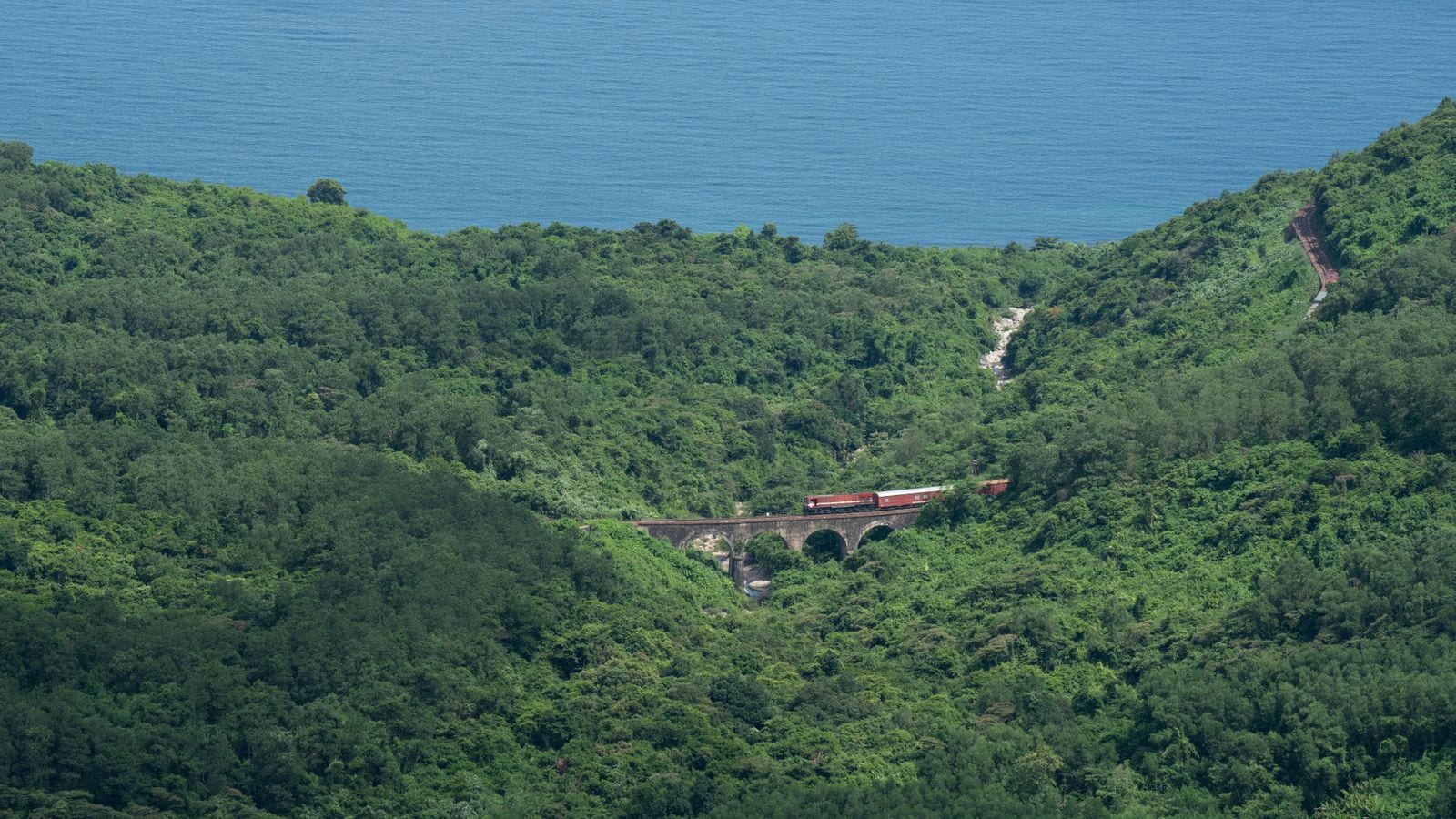 A short train crossing a stone viaduct on a jungle hillside, the open sea filling the top of the frame above the ridge