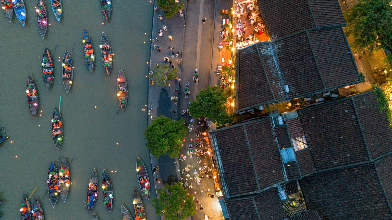 An old town waterfront straight from above at dusk: tiled roofs on one side, a quay of lit stalls, and the river below crowded with boats carrying paper lanterns