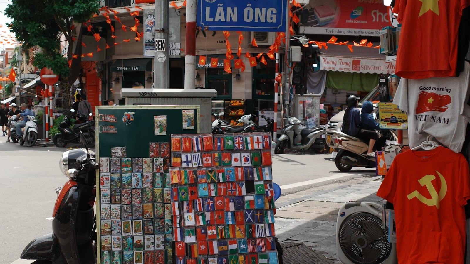 A pavement souvenir stall of flag patches and fridge magnets under the Lan Ong street sign in Hanoi, with scooters passing behind