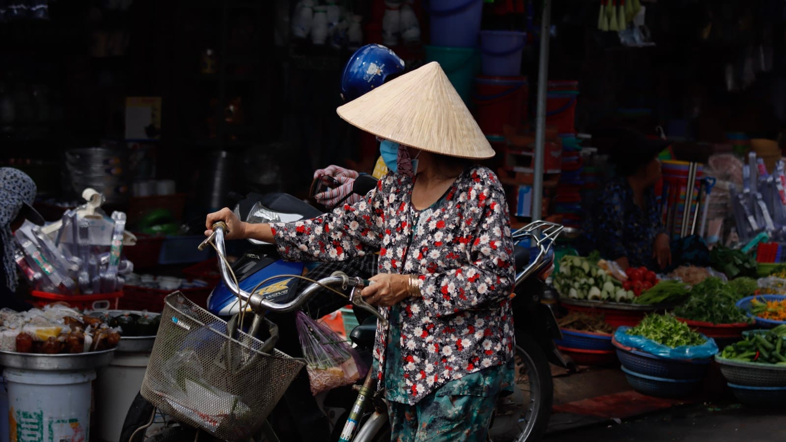 A woman in a conical hat and a long-sleeved floral shirt pushing a bicycle past market baskets of vegetables